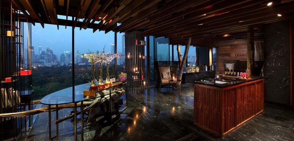 A modern interior of a spa or wellness center featuring sleek wooden decor, large windows showcasing a city view, and a reception area with candles and plants.