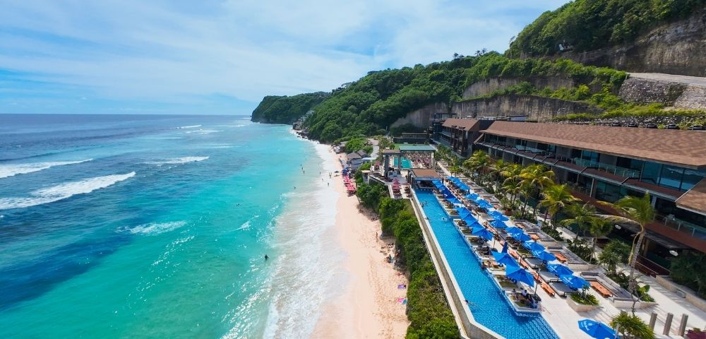 Aerial view of a resort with a bright blue infinity pool and a sandy beach with turquoise water.
