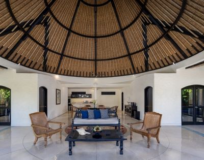 Open-air living room with a thatched domed ceiling, wicker armchairs, and a dark wood coffee table and sofa with blue cushions.