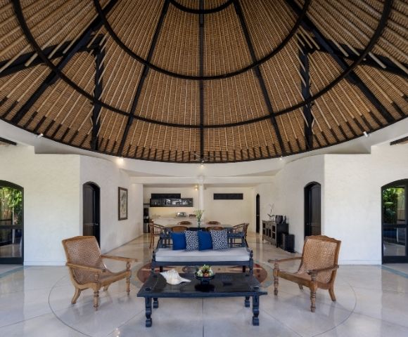 Open-air living room with a thatched domed ceiling, wicker armchairs, and a dark wood coffee table and sofa with blue cushions.