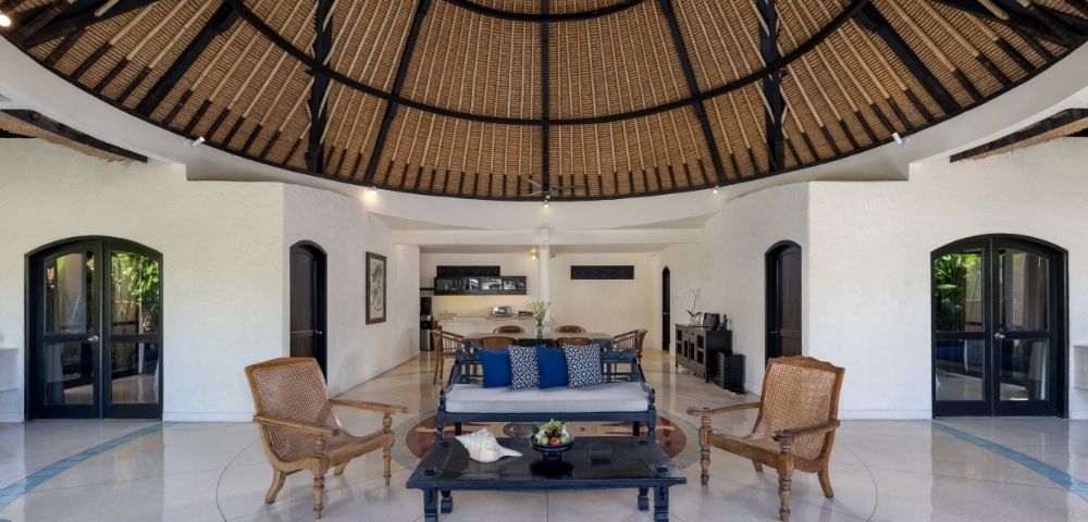 Open-air living room with a thatched domed ceiling, wicker armchairs, and a dark wood coffee table and sofa with blue cushions.
