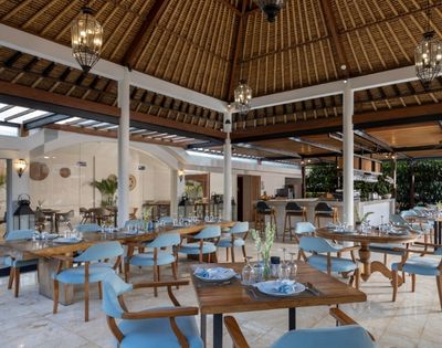 Open-air dining area with tables set for a meal, featuring wooden tables, light blue chairs, and a thatched roof structure.