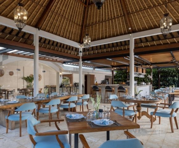 Open-air dining area with tables set for a meal, featuring wooden tables, light blue chairs, and a thatched roof structure.