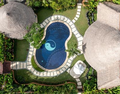 Aerial view of a dark blue swimming pool with a yin yang symbol in the center, surrounded by a stone path and lush green grass, next to buildings with thatched roofs.