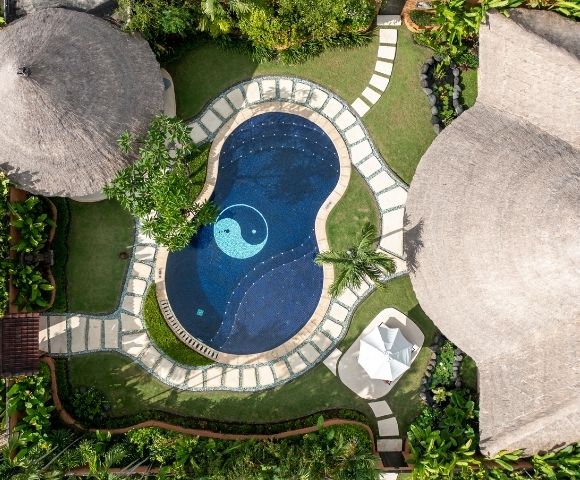 Aerial view of a dark blue swimming pool with a yin yang symbol in the center, surrounded by a stone path and lush green grass, next to buildings with thatched roofs.
