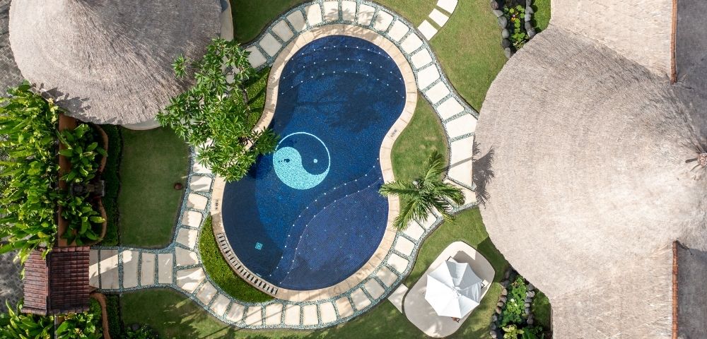 Aerial view of a dark blue swimming pool with a yin yang symbol in the center, surrounded by a stone path and lush green grass, next to buildings with thatched roofs.