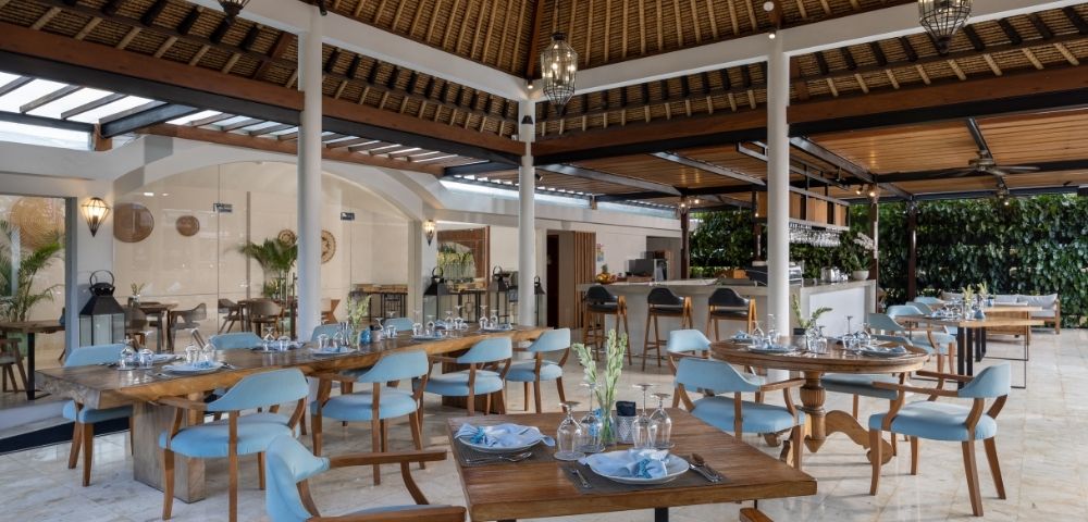 Open-air dining area with tables set for a meal, featuring wooden tables, light blue chairs, and a thatched roof structure.