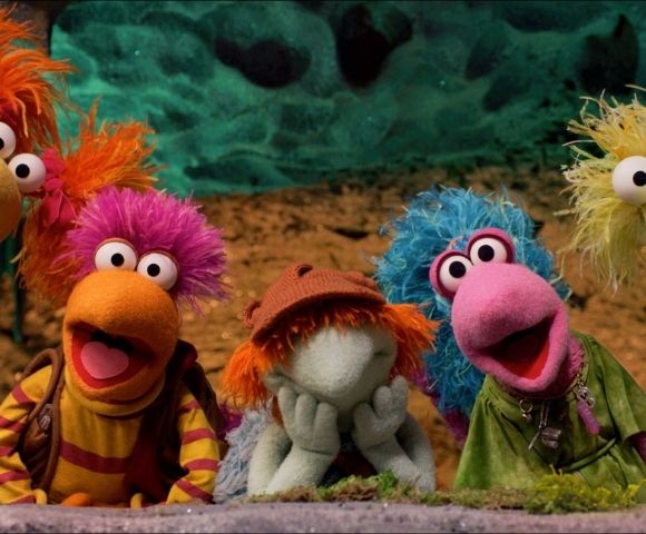 Colorful puppet characters with vibrant hair and big eyes appear against a textured, earthy background.