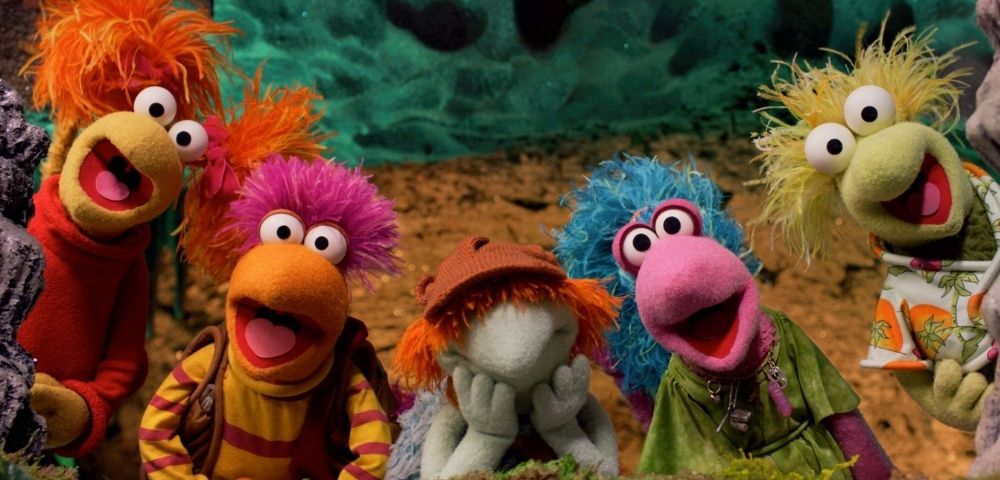 Colorful puppet characters with vibrant hair and big eyes appear against a textured, earthy background.
