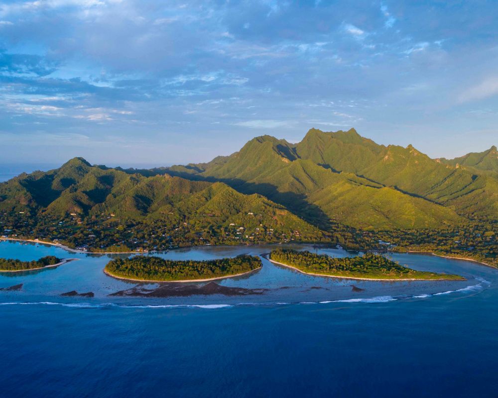 Panoramic aerial view of a lush green volcanic island with a town nestled along its coastline, fringed by a tranquil turquoise lagoon and several small, palm-covered islets.