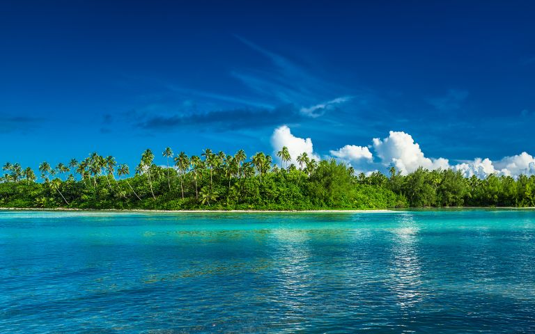 A wide panoramic view of a tropical island with lush green vegetation and palm trees, bordered by a white sandy beach and turquoise blue ocean under a clear sky with fluffy white clouds.