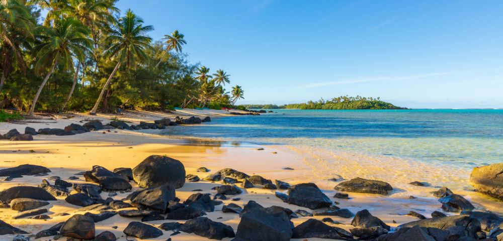 Golden sand beach lined with palm trees and dark rocks, leading to calm, clear turquoise water and a distant green island under a bright blue sky.