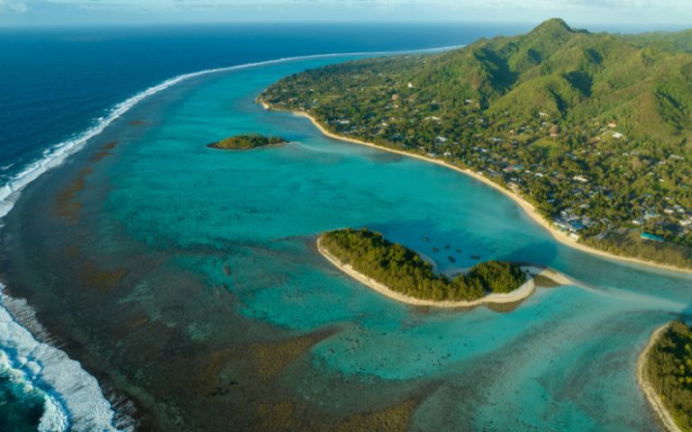 Aerial view of a tropical coastline with clear turquoise water, white sandy beaches, lush green islands, and a small village nestled amongst palm trees.