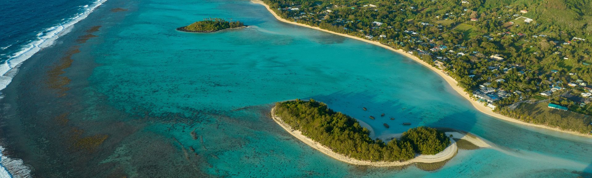 Aerial view of a tropical coastline with clear turquoise water, white sandy beaches, lush green islands, and a small village nestled amongst palm trees.