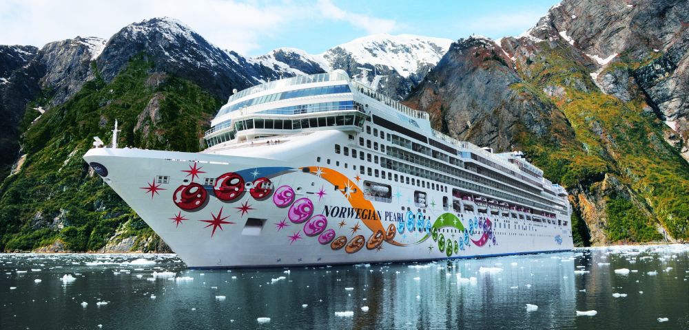 A large, white cruise ship named Norwegian Pearl, adorned with colorful bubble-like graphics and stars, sails on calm water near a steep, lush green mountainside with patches of snow.