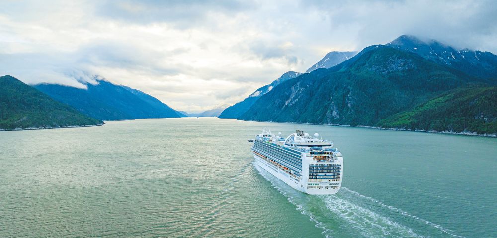 A large white cruise ship sails on calm, greenish water through a fjord with steep, tree-covered mountains on either side and a cloudy sky overhead.