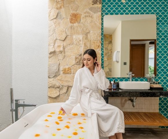 The image features a woman wearing a white robe, sitting in a bathtub filled with water and flowers. She is surrounded by a beautiful and relaxing ...