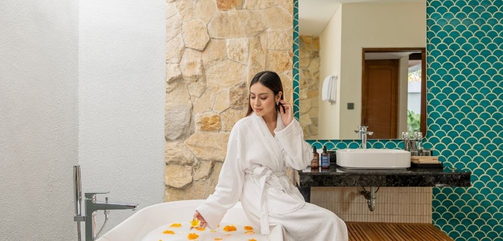 The image features a woman wearing a white robe, sitting in a bathtub filled with water and flowers. She is surrounded by a beautiful and relaxing ...