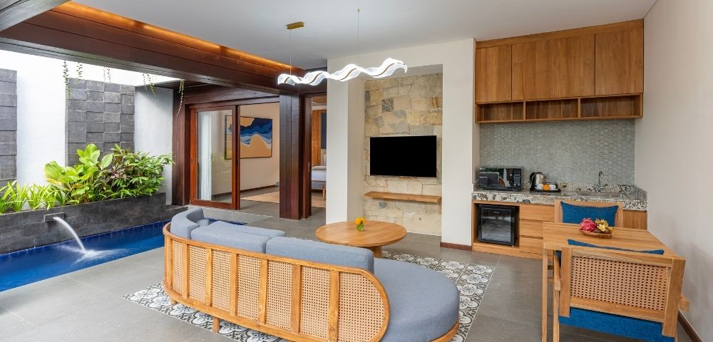 A modern living space with a curved gray sofa, a small plunge pool with a waterfall, a kitchenette, and a dining area.