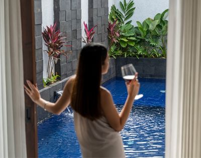 Woman in a towel holds a glass of red wine by a private pool with lush greenery.