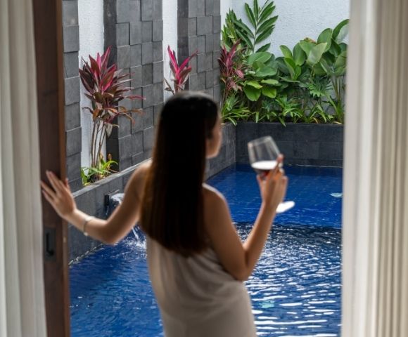 Woman in a towel holds a glass of red wine by a private pool with lush greenery.