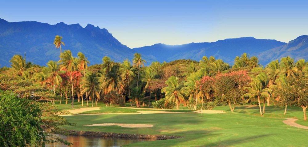 A lush green golf course fairway with sand traps and palm trees in the foreground, with a vibrant pink flowering tree and dense tropical foliage behind. In the background, dramatic blue mountains rise against a clear sky at sunrise or sunset.