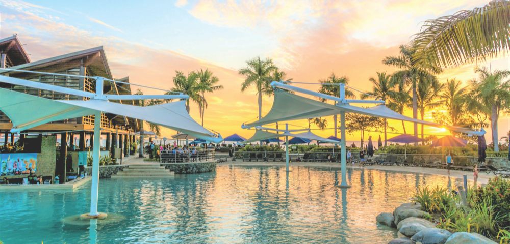 A resort swimming pool and restaurant area at sunset, with palm trees silhouetted against a vibrant orange and yellow sky, and people relaxing under large shade structures.