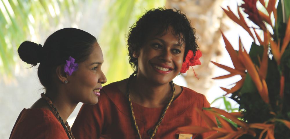 Two women in orange tops with flowers in their hair smile, with lush green foliage in the background.