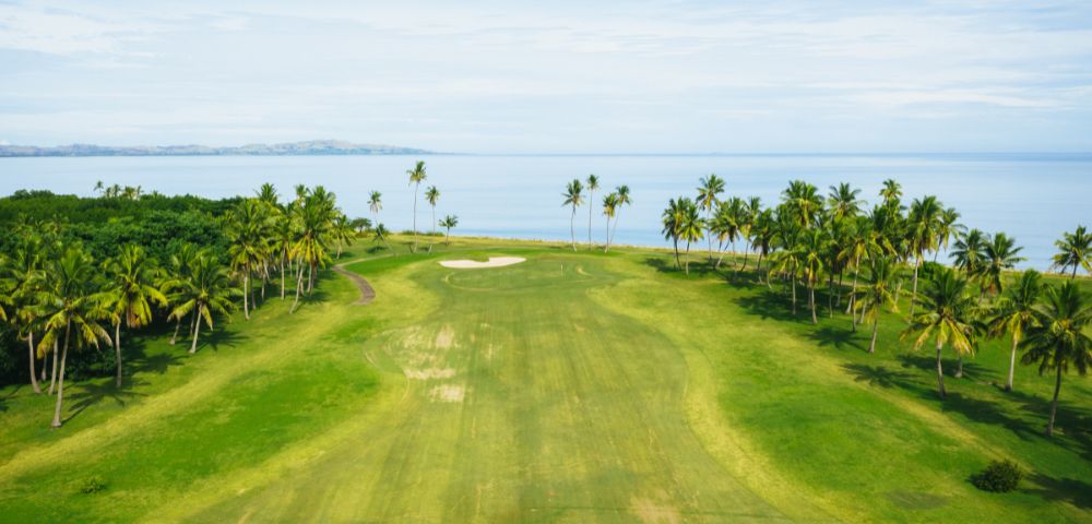 An aerial panoramic view of a lush green golf course fairways lined with palm trees, with a sand trap visible, bordering a calm blue ocean under a bright sky.