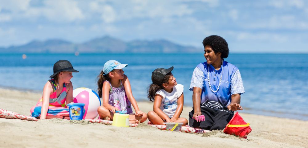 A group of four children and an adult sit on a sandy beach with the ocean and distant hills in the background. Three children are wearing hats and swimwear, with beach toys scattered around them. The adult, wearing a blue shirt and traditional jewelry, interacts with one of the children.