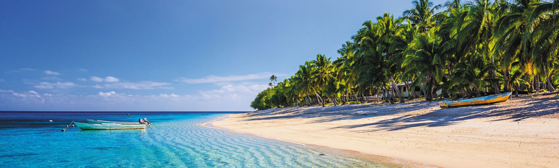 A tranquil tropical beach with clear turquoise water lapping a sandy shore lined with palm trees, featuring two boats in the shallow water and one on the sand.