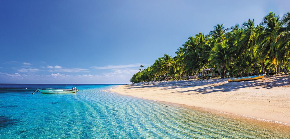 A tranquil tropical beach with clear turquoise water lapping a sandy shore lined with palm trees, featuring two boats in the shallow water and one on the sand.