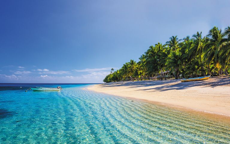 A tranquil tropical beach with clear turquoise water lapping a sandy shore lined with palm trees, featuring two boats in the shallow water and one on the sand.