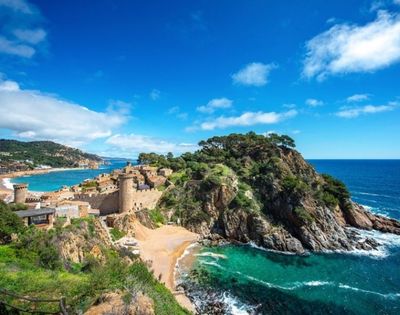 Coastal view of a historic stone castle on a rocky peninsula surrounded by turquoise sea and a small sandy beach under a bright blue sky.