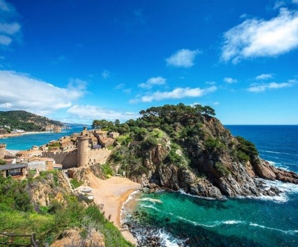 Coastal view of a historic stone castle on a rocky peninsula surrounded by turquoise sea and a small sandy beach under a bright blue sky.