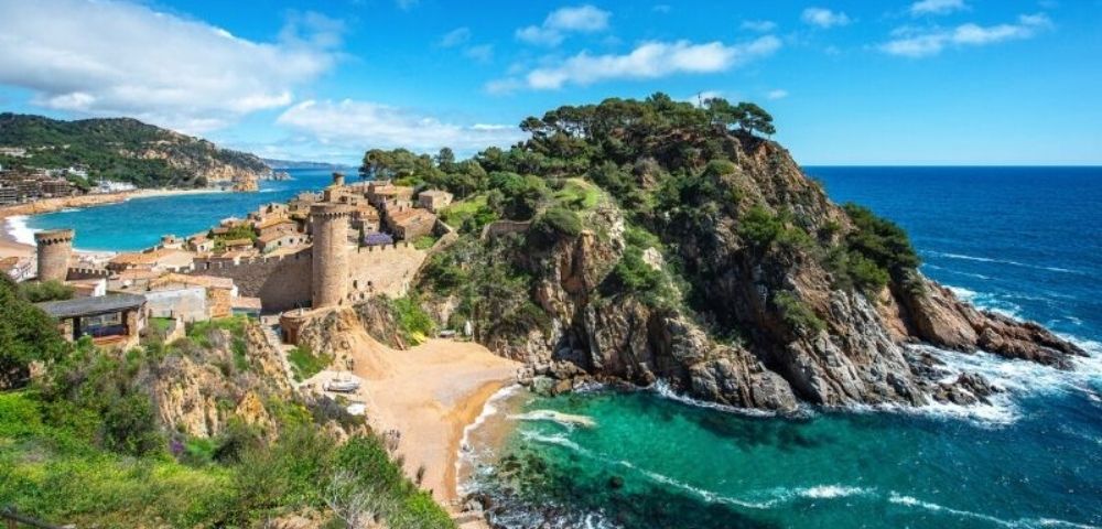 Coastal view of a historic stone castle on a rocky peninsula surrounded by turquoise sea and a small sandy beach under a bright blue sky.