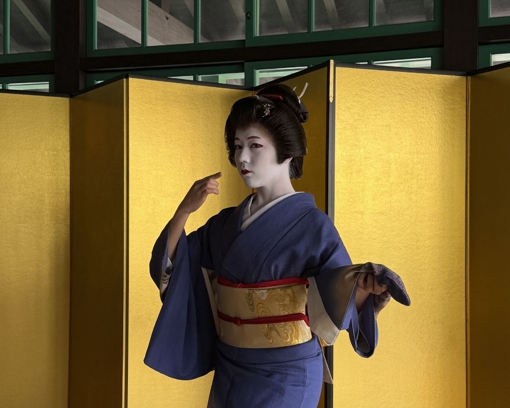 A performer in traditional Japanese white makeup and a dark blue kimono gestures with her finger in front of a gold screen.