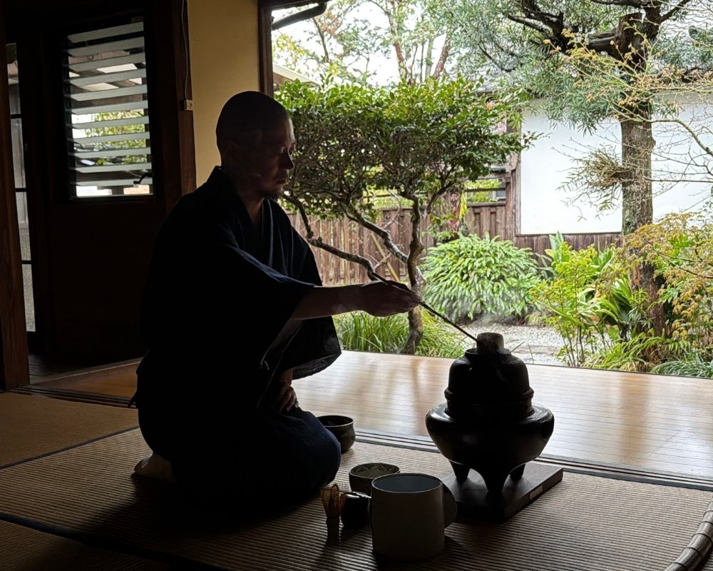 A person in a dark robe kneels on a tatami mat, preparing matcha tea in a traditional Japanese tea ceremony.