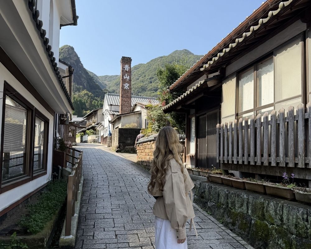 A person with long blonde hair walks down a narrow, cobblestone street lined with traditional Japanese buildings and a wooden fence.