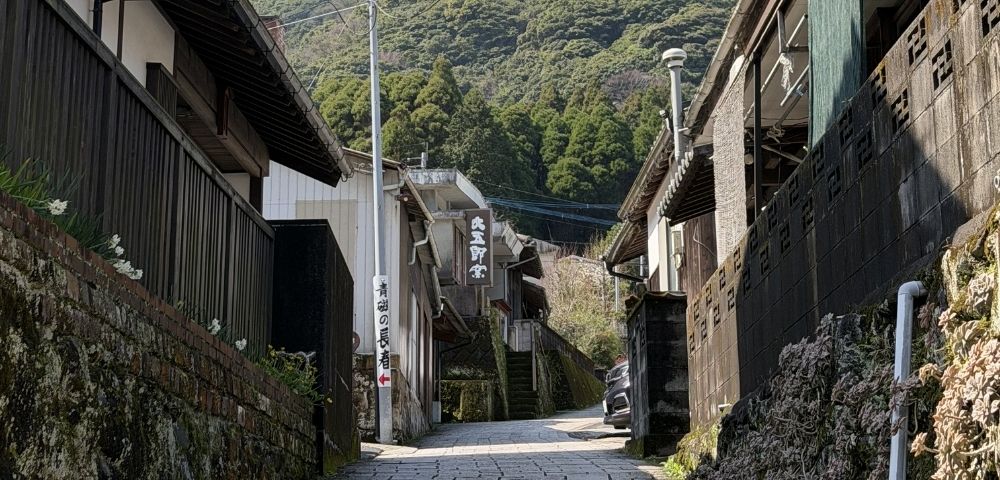 Narrow alleyway with traditional Japanese buildings and lush green trees in the background.