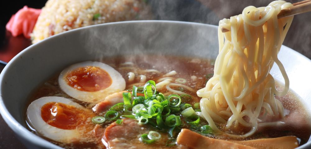 A bowl of steaming ramen with two halves of a soft-boiled egg, sliced green onions, and chashu pork, with a portion of fried rice in the background.