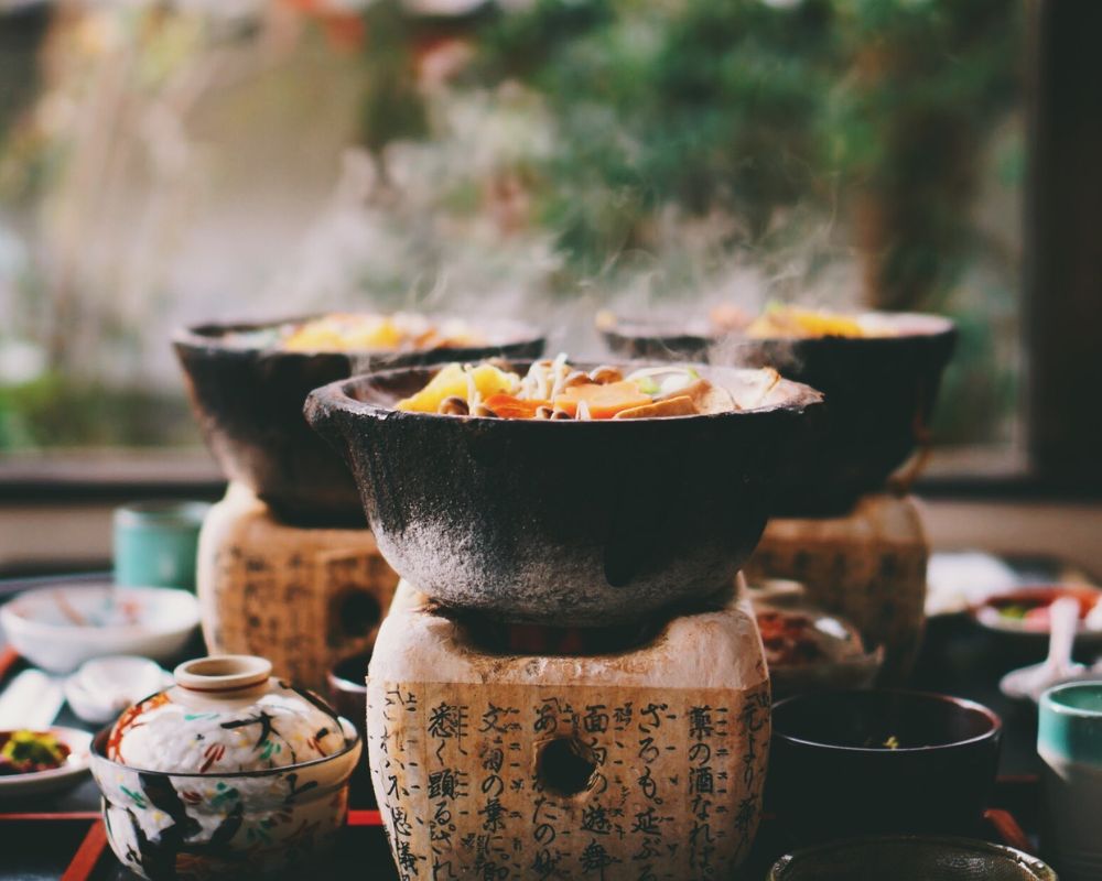 Several stone bowls filled with hot, steaming food sit on decorative wooden stands, set within a traditional Japanese meal presentation with various ceramic dishes.