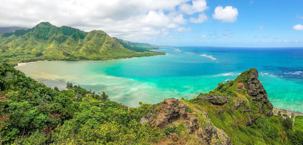 Panoramic view of a lush green coastline meeting turquoise waters, with a dramatic rocky peak in the foreground and verdant, mountainous terrain stretching into the distance.