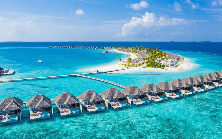 Aerial view of an overwater bungalow resort in the Maldives with thatched-roof villas on stilts above clear turquoise water and a white sand island with palm trees.