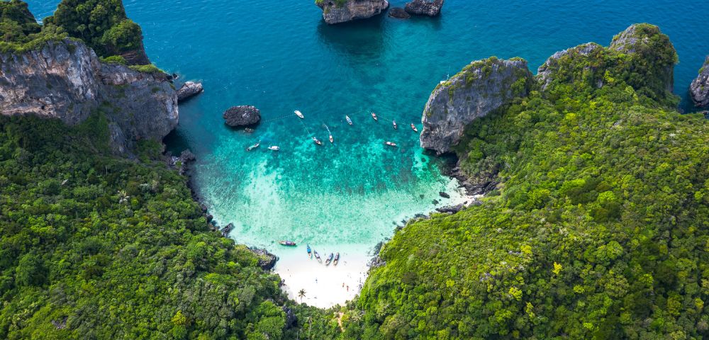 Aerial view of a secluded tropical cove with a white sandy beach, turquoise clear water, lush green cliffs, and several longtail boats anchored offshore.