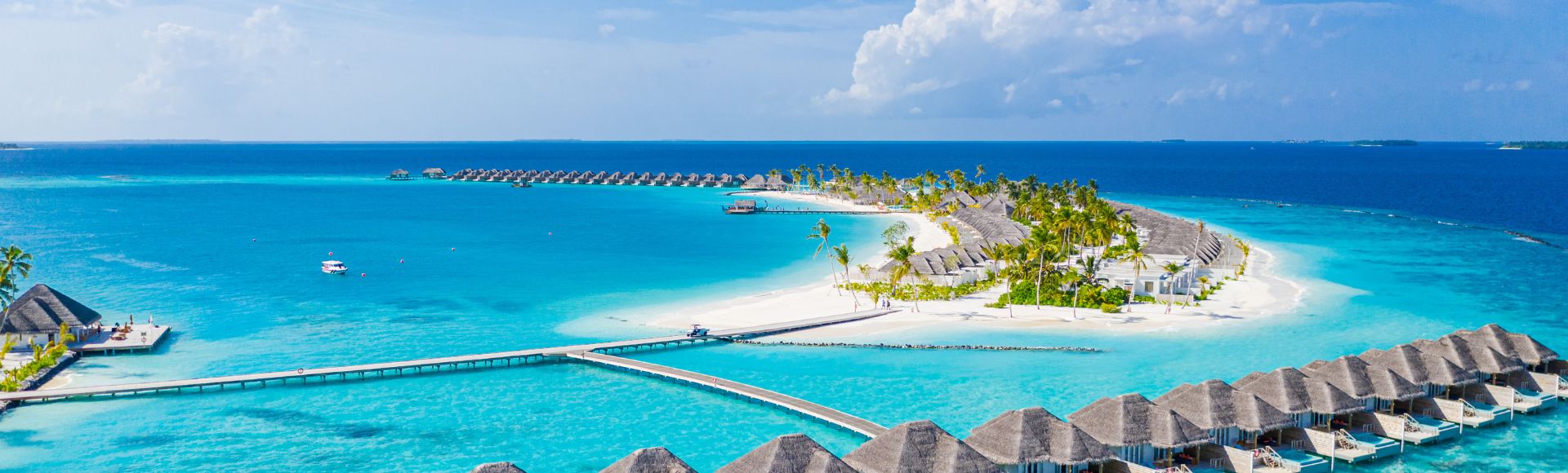 Aerial view of a tropical island resort with overwater bungalows and villas on pristine white sand beaches, surrounded by vibrant turquoise waters under a bright blue sky.
