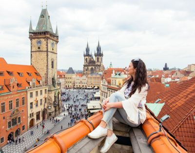 A woman sits on a rooftop overlooking a historic European city square with iconic clock towers and church spires.
