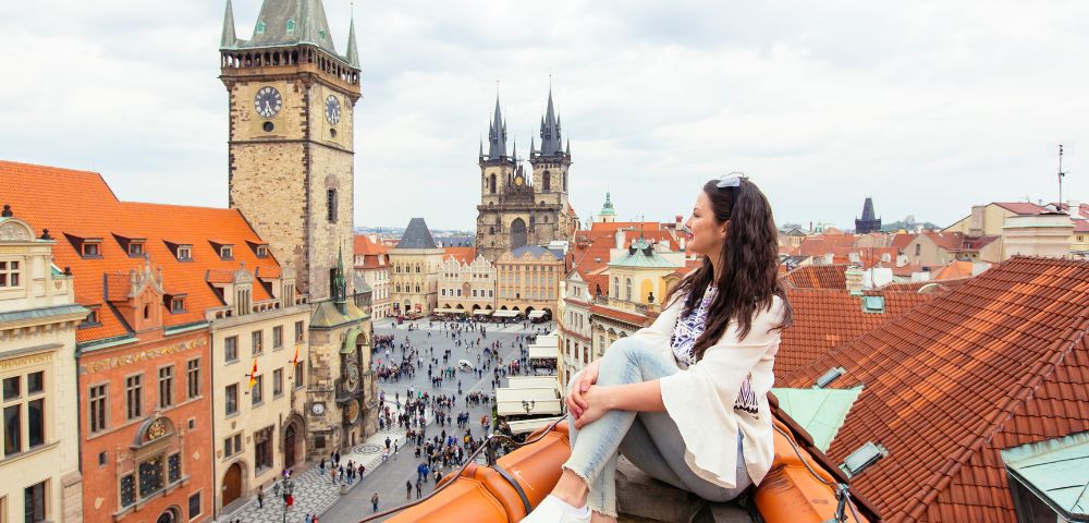 A woman sits on a rooftop overlooking a historic European city square with iconic clock towers and church spires.