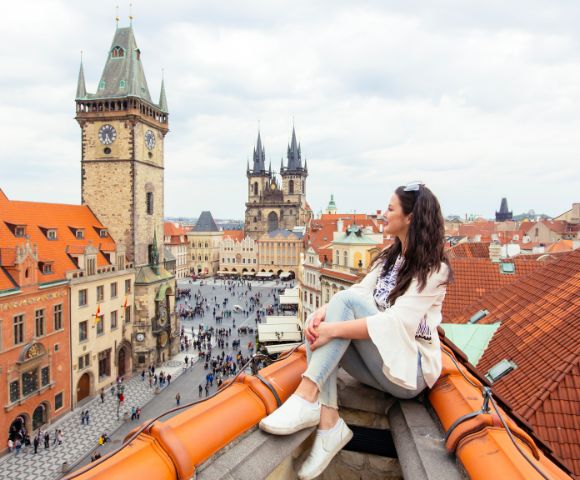 A woman sits on a rooftop overlooking a historic European city square with iconic clock towers and church spires.