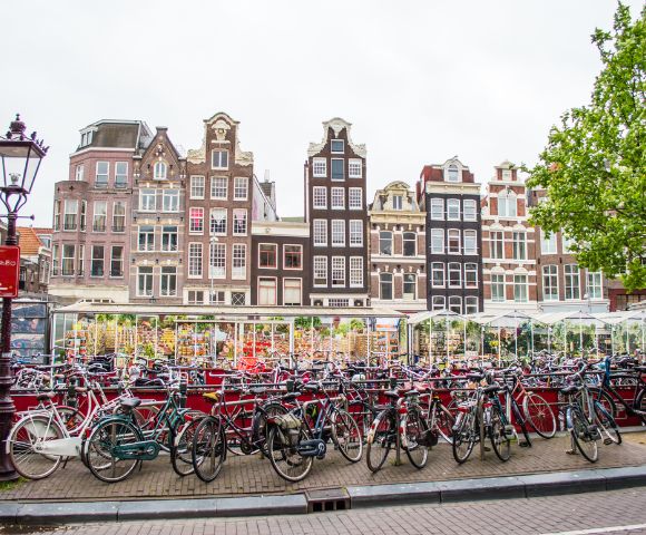 A large outdoor bicycle parking lot is in the foreground, with numerous bikes lined up. Behind the bikes are several glass-enclosed market stalls filled with goods, and beyond that are tall, narrow historic buildings with distinct gabled roofs under a cloudy sky.